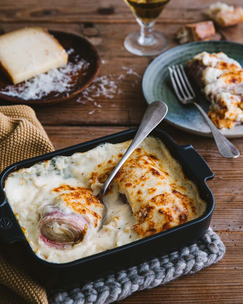 Plat à gratin avec des endives roulées au jambon avec de la sauce béchamel et une assiette avec une endive dedans. Un plat parfait pour l